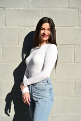 Vertical Caucasian portrait of a pretty brunette girl in a white blouse and blue jeans stands near the wall of a building on a spring day in the city. Model is happy to pose.