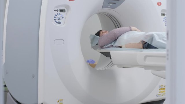 Woman lying on the CT or MRI scanner during machine imaging her body, lights up infrared rays and female patient passes through the circle, crane shot from down to up, room interior, active scene.