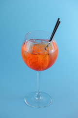 Cold summer alcohol cocktail with ice and orange in a wine glass and black plastic straw on light pastel blue background