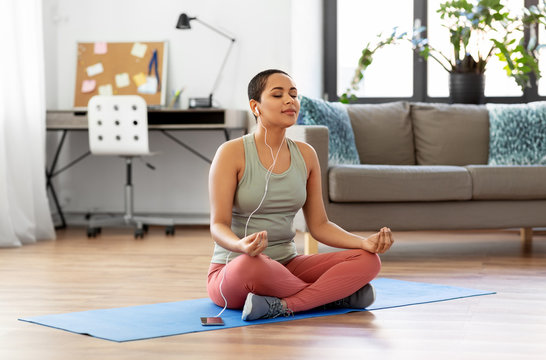 mindfulness, spirituality and healthy lifestyle concept - african american woman in earphones listening to music on smartphone and meditating in lotus pose at yoga studio