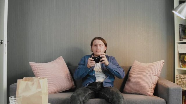 Young gamer holding joystick and playing video game sit alone on couch at home