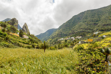 Obraz premium Roques de Pedro and Petra behind a beautiful palm tree seen from the valley in La Hermigua on La Gomera. April 15, 2019. La Gomera, Santa Cruz de Tenerife Spain Africa. Travel Tourism