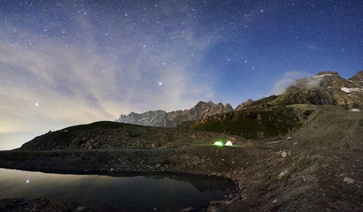 Fototapeta na wymiar Magnificent view of majestic rocky hills with mountain lake and illuminated tourist camp tens in mountain valley under fantastic starry sky. Concept of travelling, camping in Alps and nature.