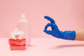 Woman's hand in rubber glove with sponges and a bottle of dishwashing liquid on the pink background.