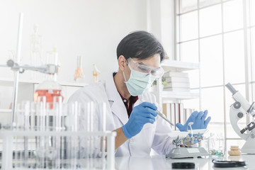 A male scientist with black hair wearing white coat and protective glassware and face mask working...