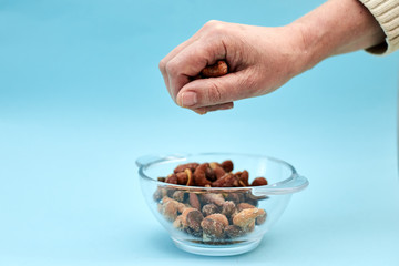 A hand is holding a handful of nuts over the glass bowl with nuts on light blue background