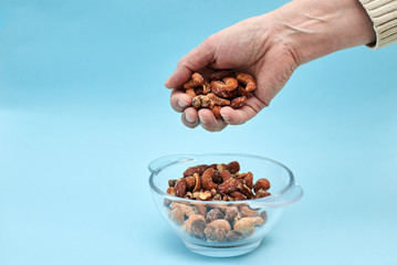 A hand is holding a handful of nuts over the glass bowl with nuts on light blue background