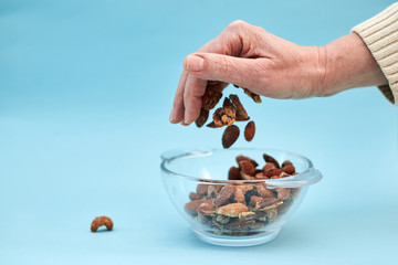 A hand is throwing nuts into the glass bowl with nuts, couple of nuts are laying aside, on light blue background