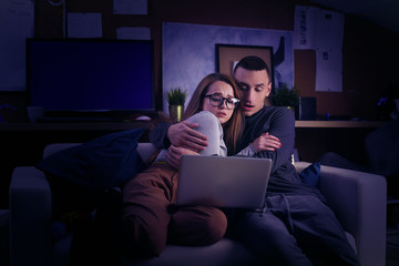 A couple is sitting on a couch and looking at a computer. Couple watching a movie, series, horror together. The girl was frightened and leaned against the man. The couple is watching horror movies.