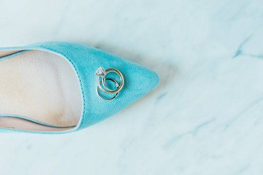 A baby blue wedding slipper with two wedding rings on top of a marble surface