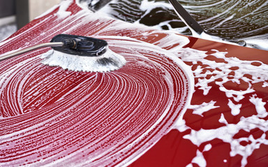 Red car washed in self serve car wash, brush leaving strokes in white soap foam as front hood is...