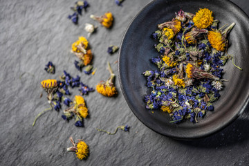 mixture of dry flowers for herbal tea styled studio shot on a black stone background in a black ceramic dish 