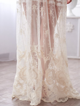 Close-up picture of the bottom of beautiful white lace peignoir night gown on young woman, standing in the middle of light bedroom with white furniture.