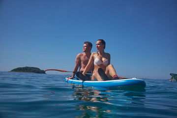 Young and beautiful couple of lovers sitting on a surfboard in the open ocean on a background of sky.