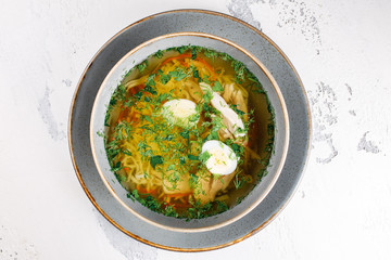 soup with quail egg, noodles, chicken and herbs in a gray plate close-up on a white texture background. top view.