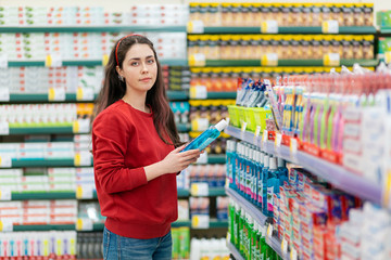 A young Caucasian woman in a red sweater holds a bottle of shampoo and smiles tenderly. In the background, shelves with cosmetics blurred. The concept of buying cosmetics