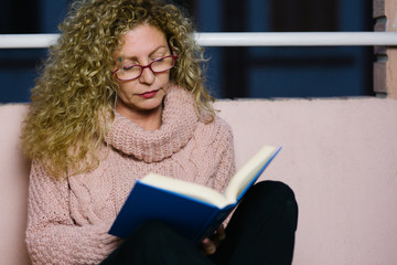a mature woman with glasses sitting on the balcony floor reading a book during the quarantine of the covid19