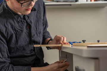 a teenager with glasses and a dressing gown is sitting at a desk engaged in carpentry. activities at home during the period of quarantine and self-isolation