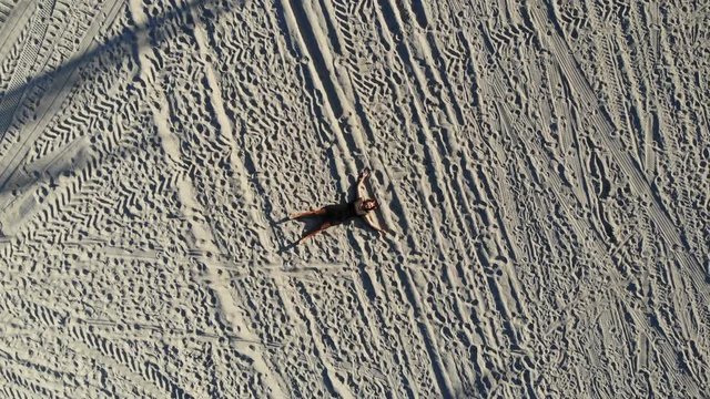 Woman lying starfish on the sand as drone shot pulls up from her.