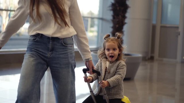 Airport terminal, young unrecognizable mother riding her cute daughter on a small yellow suitcase. Mom with yellow suitcase and daughter are having fun before their departure. Close up