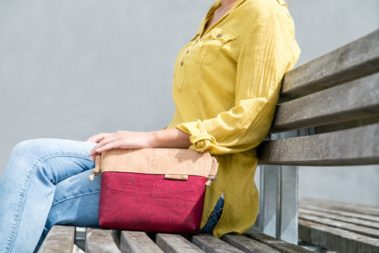 Close Up Of Young Woman Sitting On Wooden Bench In City With Her Eco Friendly Cork Bag From The Side. Enjoying Warm Sunny Day