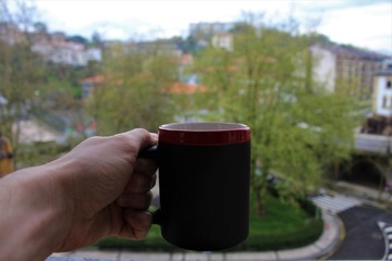 
hand holding red and black cup of coffee by the window.