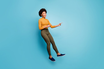 Fototapeta na wymiar Full size photo of positive dark skin girl jump help choose decide advise feedback point index finger copyspace wear yellow striped sweater green pants trousers isolated blue color background