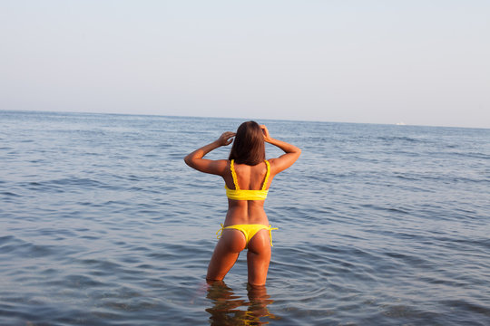 Beautiful tanned woman in yellow swimsuit stands in the sea bathing on the beach