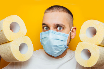 A handsome guy in a protective mask hides behind a wall of toilet paper to prevent coronavirus infection. Yellow background. Flash concept COVID-19. Home insulation.
