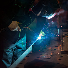 Worker welding a section of metal square tube with a welding torch creating sparks and fumes on the work bench of a factory workshop