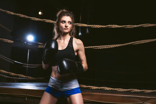 Young girl in sportswear: a t-shirt and shorts wearing boxing gloves. Athletic woman posing on a background of a boxing ring in the dark gym. Power and motivation concept