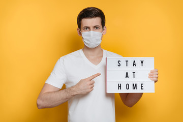 Young serious man in medical mask pointing finger at board with Stay home text. Isolated on yellow background. Stay at home social media campaign for coronavirus or COVID-19  prevention 