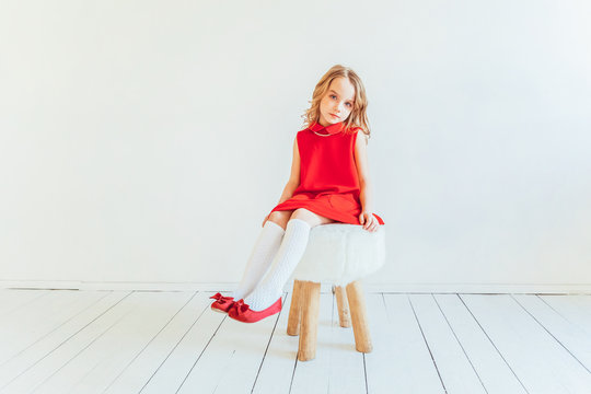 Stay Home Stay Safe. Sweet Little Girl In Red Dress Sitting On Chair Against White Wall At Home, Relaxing In White Bright Living Room Indoors. Childhood Schoolchildren Youth Relax Concept.