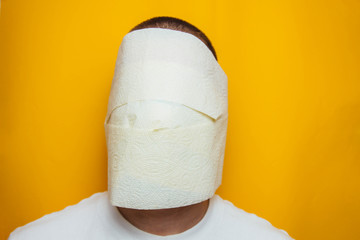 A man in a blue shirt wrapped his face in toilet paper, trying to protect himself from the coronavirus. In the studio on a yellow background. Protecting health from viruses.
