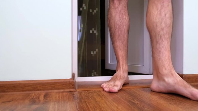 Unsexy man with hairy legs in shorts barefoot crosses the threshold, enters the balcony of a Flat, scratches his leg. Dolly Shot, Moving Camera.