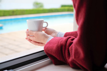 A caucasian woman drinking a cup of 
coffee by the window wearing pajamas and a red robe at home during self isolation
