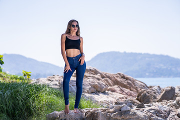 Fashion shooting with female model stand on beach stone in tropical place, behind sea, dark blue jeans, black top, sunglasses, summer time.
