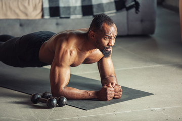 African-american man teaching at home online courses of fitness, aerobic, sporty lifestyle while being quarantine. Getting active while isolated, wellness, movement concept. Training, plank.