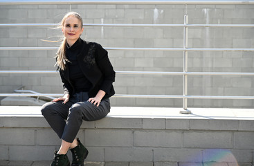 Photo of a pretty blonde Caucasian woman in a city outdoors in the afternoon on the background of the railing of the stairs. Fashionable model posing with a smile and pleasure.