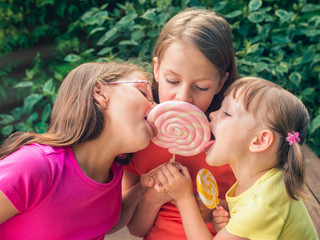 Three funny little girls in colorful T-shirts licking large lollipops - on the garden terrace