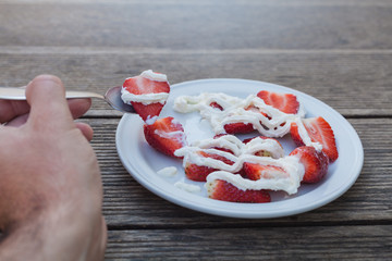 Eating strawberries with cream, hand holding spoon to eat dessert fruit on wooden table as background