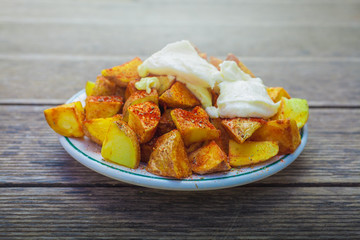 Patatas bravas o patatas gajo con salsa all i oli y pimentón, recién hechas con humo saliendo en plato blanco y sobre mesa de madera. Comida típica Valenciana y Española.