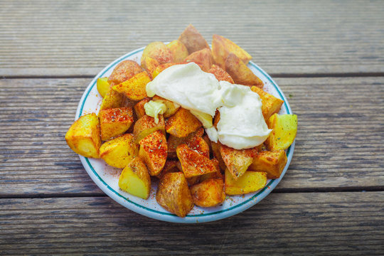 Patatas bravas o patatas gajo con salsa all i oli reci&eacute;n hechas con  humo saliendo en un plato blanco y sobre mesa de madera. Comida t&iacute;pica Valenciana y Espa&ntilde;ola.