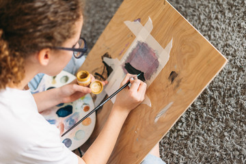 Curly hair girl teenager in glasses in white t-shirt sitting on floor painting on wooden desk at home