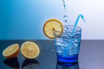 A mug of ice and water sits on a mirrored table with sliced lemons.Preparation of a fresh drink tonic,juice or cocktail.Water splash,water drop.Splashes of water fall into a glass with lemon and ice