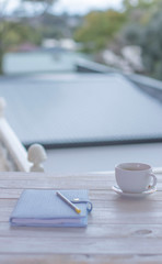 Notebook, pen and coffee cup on a wooden table on balcony. Blurred town house background