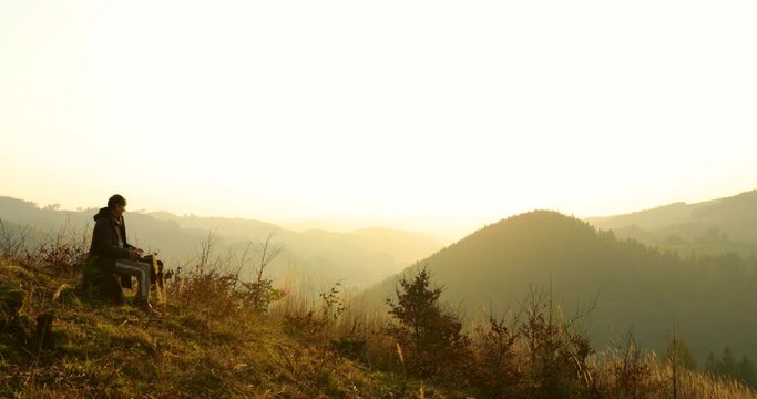 Old woman sitting on a dead tree stump and watching an orange sunset on a mountainous and hilly landscape when the sun sets over one mountain peak in the distance captured at 4k 60fps