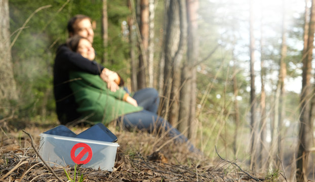 Digital detox concept. Couple spend time together in nature without smartphones. Man and woman sitting in park or forest, hugging, phones in box with prohibiting sign on foreground. Banner.