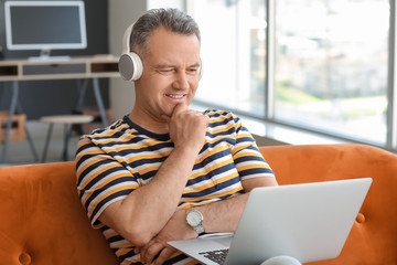 Mature man with laptop and headphones sitting on sofa at home - obrazy, fototapety, plakaty