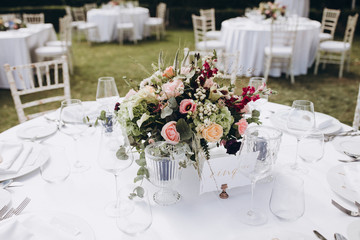 Wedding banquet. Tables in a green meadow are decorated with flower arrangements, on the tables are white tablecloths, plates with napkins, glasses and candles, cutlery. White bulbs hang over tables